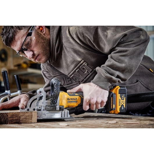 A person using a DEWALT DCW682P2 cordless biscuit joiner tool to work on a piece of wood, showing the tool in use on a workbench. The person is wearing a brown DEWALT sweatshirt and there are wood clamps and dust visible in the workspace.