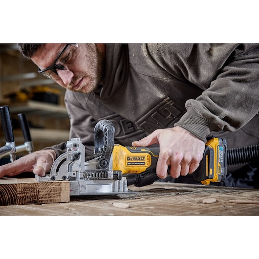 A person using a DEWALT DCW682P2 cordless joiner tool to cut into a piece of wood on a workbench, with clamps and dust extraction attached.