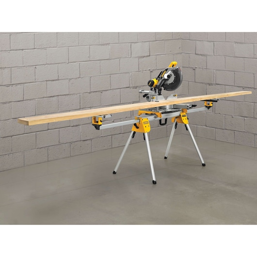 A DEWALT miter saw mounted on a DE7033 portable saw stand, holding a long piece of lumber in a workshop with gray brick walls and a concrete floor.