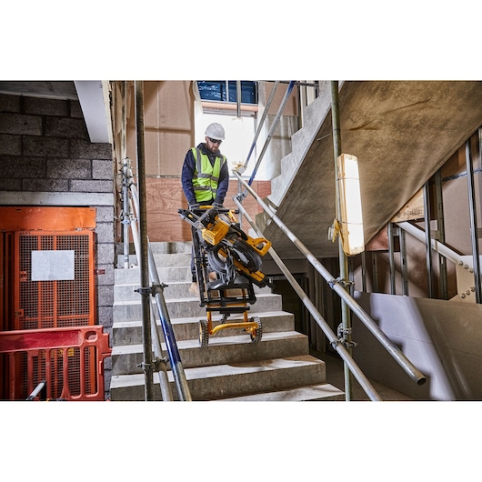 A worker in a safety vest is transporting a yellow DEWALT tool stand with attached saw up a concrete staircase at a construction site.