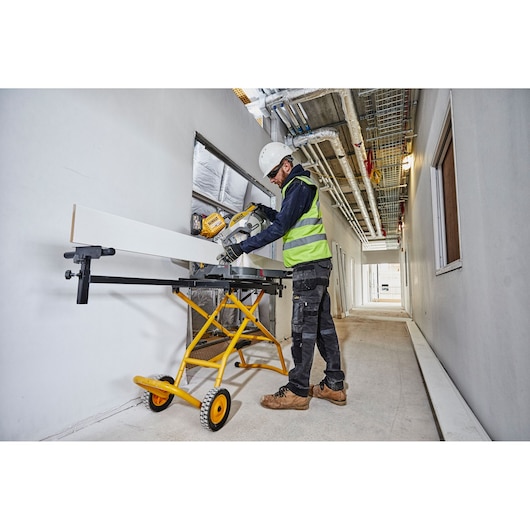 A worker wearing safety gear cuts a long white board with a sliding mitre saw mounted on a yellow DEWALT stand (model DE7260) in a construction hallway.