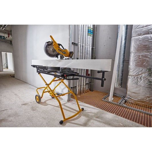 A DEWALT mitre saw mounted on a yellow mobile stand is set up indoors at a construction site, with a large white board positioned on the stand for cutting.