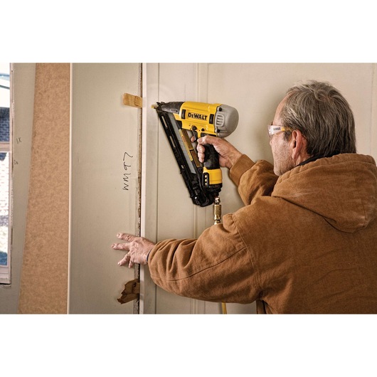 A person using a DEWALT pneumatic nailer to attach trim to a doorway during a construction or renovation project. The nailer has a yellow and black design and is connected to an air hose.