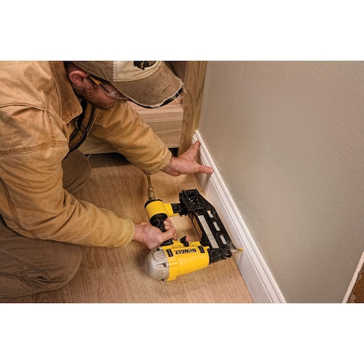 A person using a DEWALT nail gun to attach a baseboard to the wall, kneeling on a wooden floor. The nail gun is yellow and connected to an air hose.