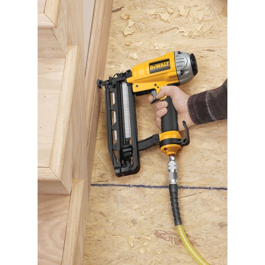 A DEWALT pneumatic nail gun (model DPN1664) being used to install wooden stair trim on an unfinished plywood surface. The nail gun is connected to an air hose.