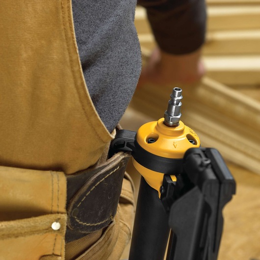 Close-up view of a DEWALT tool attached to a worker's tool belt. The worker is wearing a brown apron and is in a wood workshop environment.