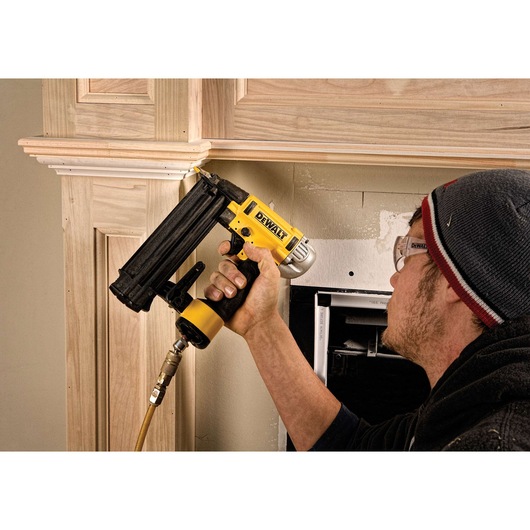 A person using a DEWALT pneumatic nailer to install wooden trim on a wall. The person is wearing safety glasses and a beanie, and the tool is connected to an air hose.