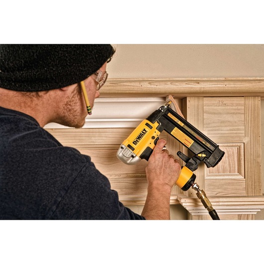 A person wearing safety glasses is using a DEWALT pneumatic nailer to install trim on wooden paneling. The tool is yellow and black, connected by an air hose.