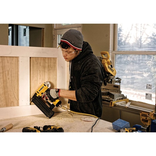 A person using a DEWALT pneumatic nailer (SKU: DPN1850PP) to secure wood panels in a workshop. Other DEWALT tools and woodworking equipment are visible on the table and in the background.