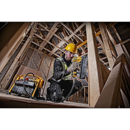 A person wearing a yellow hard hat and safety vest is working on a wooden construction site, using a DEWALT pneumatic nail gun connected to a DEWALT air compressor. The person is crouched among wooden beams, and their face is blurred.