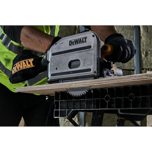 A person wearing black gloves and a yellow safety vest is using a DEWALT track saw to cut a piece of wood, with the saw blade visible beneath the material.