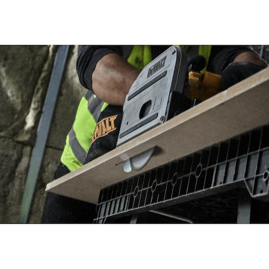 A DEWALT power tool is being used to cut a wooden board, with a close-up view of the tool and the work surface. The person operating the tool is wearing a high-visibility vest and gloves.