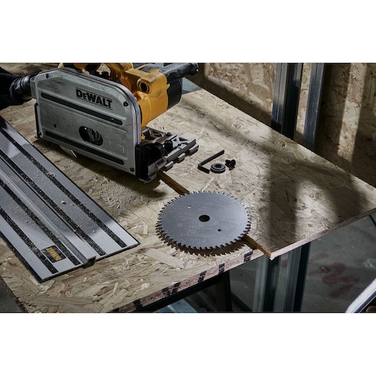 A DEWALT track saw placed on a wooden workbench, alongside a metal saw blade, track guide, and several small accessories. The setting has an industrial background with wooden panels and metal framework.