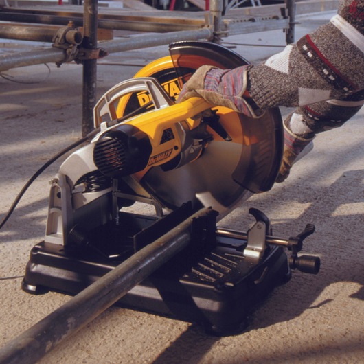A DEWALT DT1921 power saw is being used to cut a metal pipe. The saw is positioned on a portable stand and operated by a person wearing gloves and a patterned sweater. The scene is outdoors at a construction site.