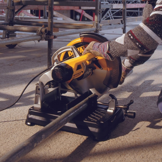 A person wearing work gloves is using a DEWALT power saw to cut a metal pipe at a construction site. The tool is plugged in and set on a solid surface, surrounded by scaffolding and construction materials.