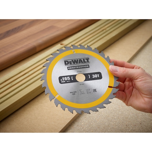 Hand holding a DEWALT DT1935 circular saw blade, 165mm diameter, 30 teeth, with wood in background.