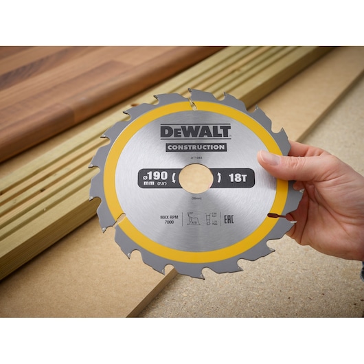Hand holding a DEWALT DT1943 circular saw blade, 190 mm, 18 teeth, with wood boards in the background.