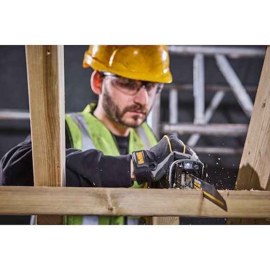 A person wearing a yellow hard hat and a green safety vest is using a DEWALT hand saw to cut a wooden beam. Sawdust is visible as the tool cuts through the wood. The person's face is obscured for privacy.