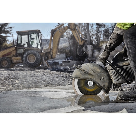 A person operating a large handheld power saw to cut through wet concrete at a construction site, with a blurred excavator and construction equipment in the background.