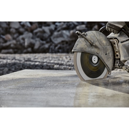 A close-up view of a circular saw cutting through concrete, with water spraying from the cutting area. The image focuses on the blade and part of the saw, showing construction work in progress.