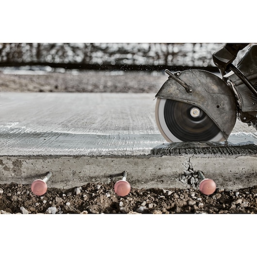 A close-up of a circular saw cutting through a concrete slab at a construction site, with nails visible in the concrete and soil below.