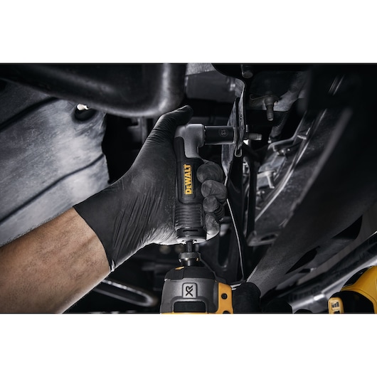 A person wearing a black glove uses a DEWALT right angle drill attachment to work on machinery in a confined space.
