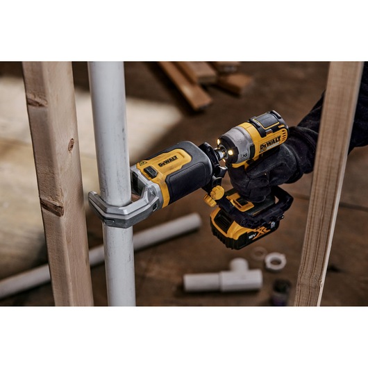 A DEWALT DT20560 power tool being used to attach a clamp to a metal pipe, held by a person wearing a glove. The tool is yellow and black, and other pipes and wooden planks are visible in the background.