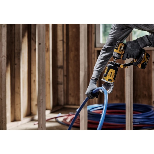 A person wearing gloves uses a DEWALT DT20560 power tool to cut a blue plastic pipe in a wooden-framed construction interior.