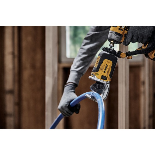 A person wearing gloves uses a DEWALT DT20560 power tool to cut a blue plastic pipe in a construction setting.