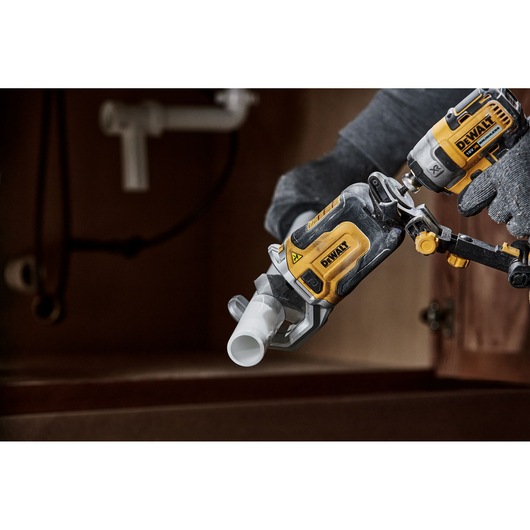 A close-up of a person using a DEWALT power tool, specifically the DT20560 model, to drill into a white plastic pipe under a sink. The person is wearing gloves and the tool is part of a plumbing installation or repair.