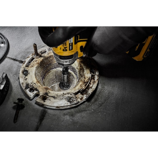 A close-up view of a DEWALT power tool being used to drill or fasten inside a metal fixture on a rough, industrial surface. The operator is wearing gloves.
