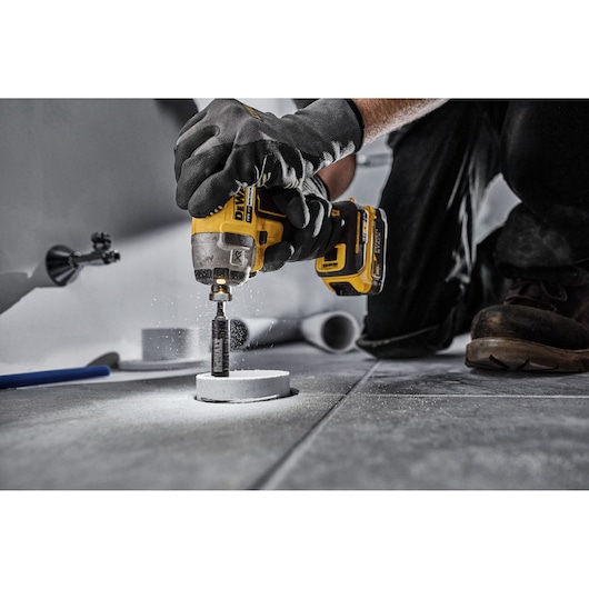 A person wearing work gloves uses a DEWALT cordless power drill to cut a circular hole in a tiled floor. Dust is visibly scattering from the drill bit.