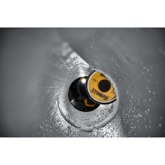 Close-up view looking through a metal pipe at a DEWALT hole saw cutting tool, shown in the process of drilling through the pipe wall, with visible metal shavings.
