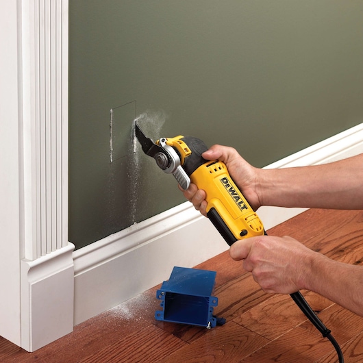 DEWALT tool cutting a hole in drywall, held by hands. Blue outlet box on floor. Dust visible near wall.