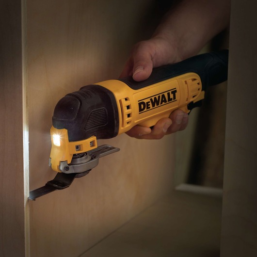 A DEWALT oscillating multi-tool being used to cut a wooden surface, held by a person's hand. The tool is equipped with a cutting blade and an LED light for enhanced visibility.