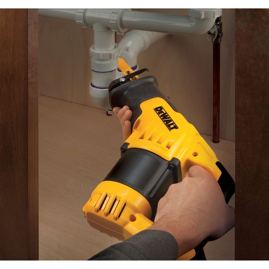 A person using a yellow and black DEWALT reciprocating saw to cut plastic plumbing pipes under a sink.