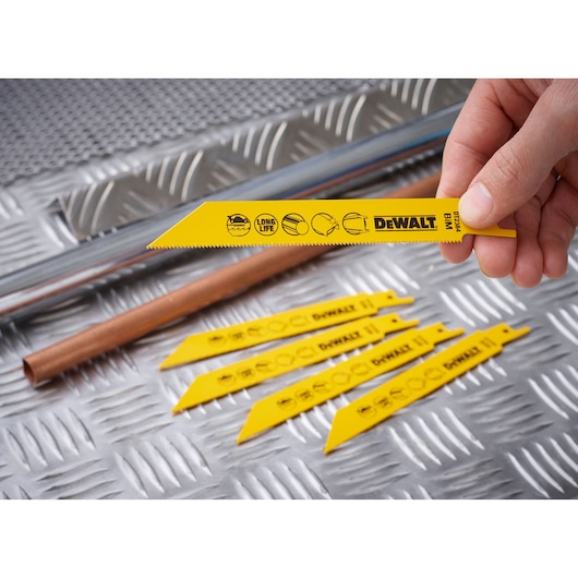 A close-up of a hand holding a yellow DEWALT reciprocating saw blade, model DT2384, with several similar blades arranged on a metal textured surface. Metal pipes are also visible in the background.