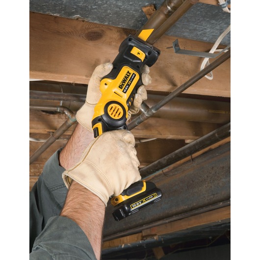 Person wearing work gloves using a DEWALT cordless reciprocating saw to cut a metal pipe near a wooden ceiling beam.