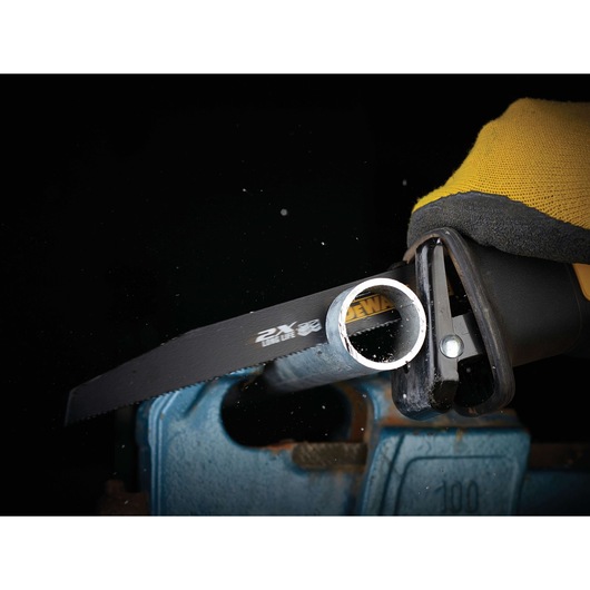 Close-up of a DEWALT reciprocating saw blade cutting through a metal pipe, held in place by a blue vise. The person operating the saw is wearing a yellow and black protective glove.