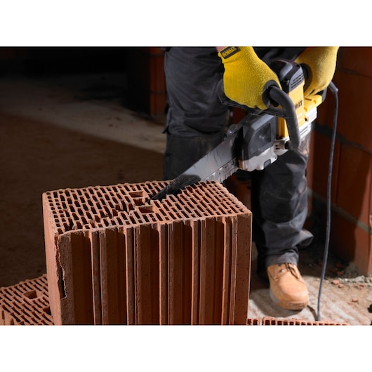 A person wearing yellow work gloves uses a DEWALT power saw to cut through a hollow construction brick. The person is standing on a construction site, and the saw is plugged in to power.
