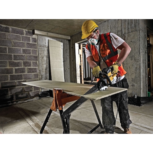 Worker uses DEWALT saw to cut a board on a worksite, wearing safety gear and protective equipment.