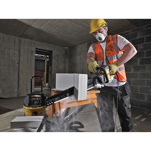 Worker uses DEWALT power tool to cut concrete block indoors, wearing safety vest, gloves, and helmet.
