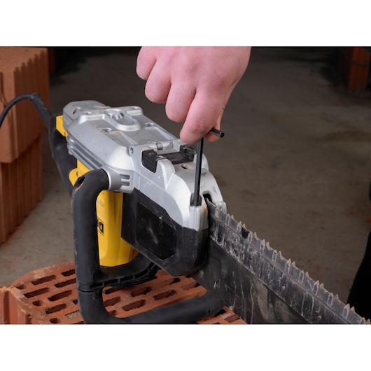 Hand uses a tool to adjust or tighten a DEWALT chainsaw blade, placed on a brick surface indoors.