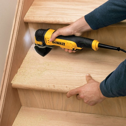 A person uses a DEWALT DT3090 power tool to sand the edge of a wooden staircase, creating wood dust. The tool is positioned