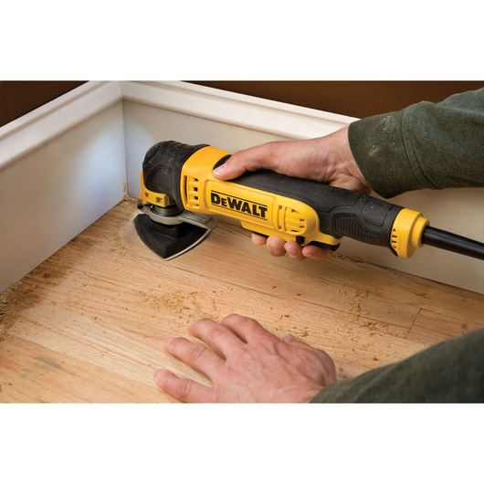 A person is using a DEWALT oscillating multi-tool to sand or work on a wooden floor near a wall and baseboard.