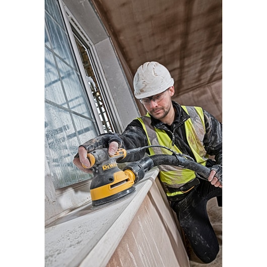 Worker uses DEWALT sander DT3102 on a window ledge. Safety gear and hi-vis vest are worn at construction site.