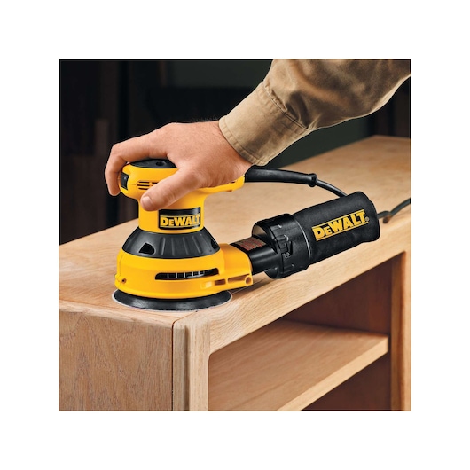 DEWALT sander in use sanding the edge of a wooden cabinet. A hand guides the yellow and black power tool.