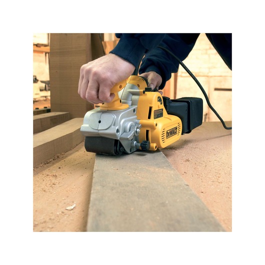 A DEWALT electric belt sander being used to smooth a piece of wood in a workshop. The tool is yellow and silver, and there is sawdust on the work surface.