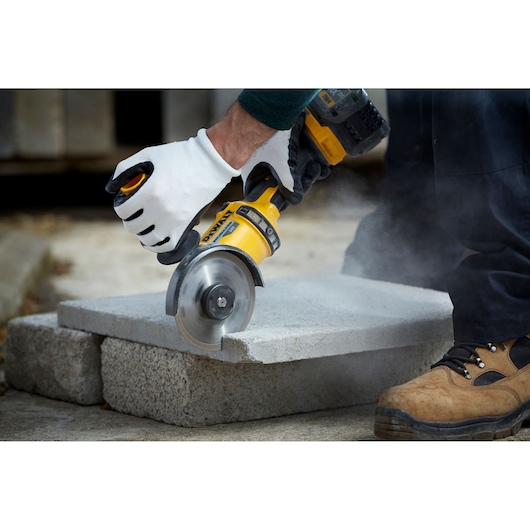 A person wearing protective gloves uses a DEWALT power tool to cut a concrete slab, with dust visible around the cutting area.