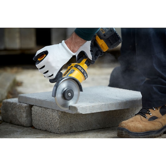 A person wearing work gloves uses a DEWALT power tool with a circular blade to cut a concrete block. The scene shows construction or masonry work, and the tool generates dust as it cuts.